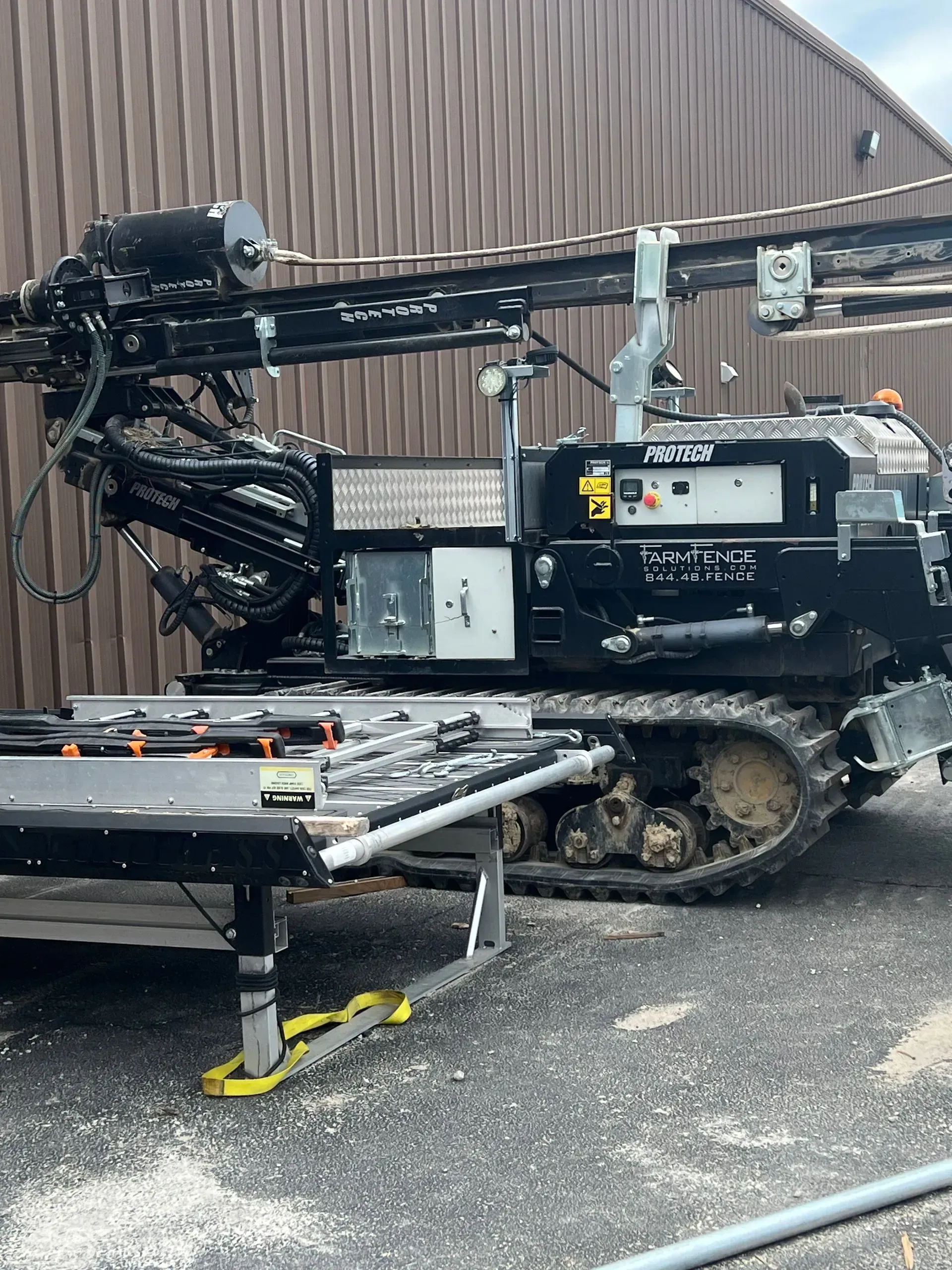 Black directional drilling machine on tracks, outdoors, by a brown building.