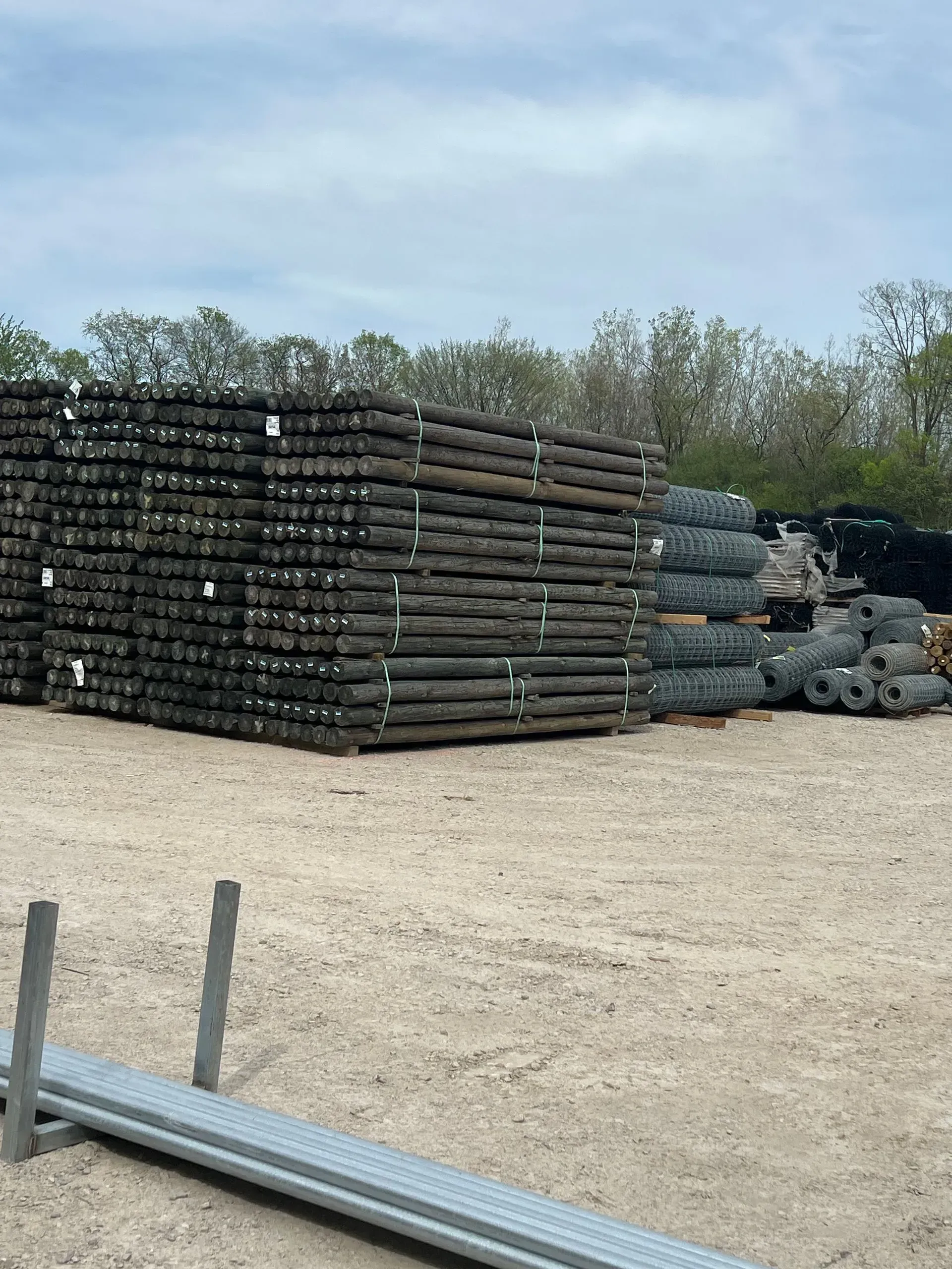 Piles of wooden fence posts and rolls of wire fencing in an outdoor storage area on a cloudy day.