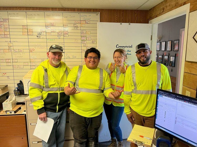 Four people in high-visibility vests pose indoors near a whiteboard and open doorway.