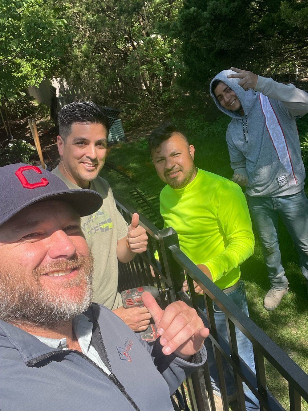 Four men posing outdoors; one takes a selfie, others give thumbs up, smile, and make a peace sign.