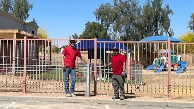 Two people in red shirts by a pink metal gate, with a playground in the background.