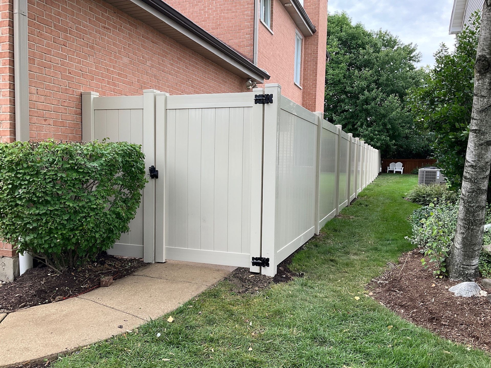 Tan vinyl fence alongside a brick building, with a gate and sidewalk.