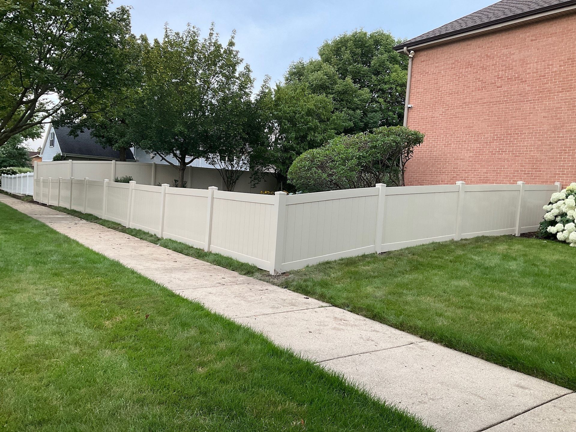 Beige vinyl fence along a sidewalk, bordering a grassy yard, near a brick building and trees.