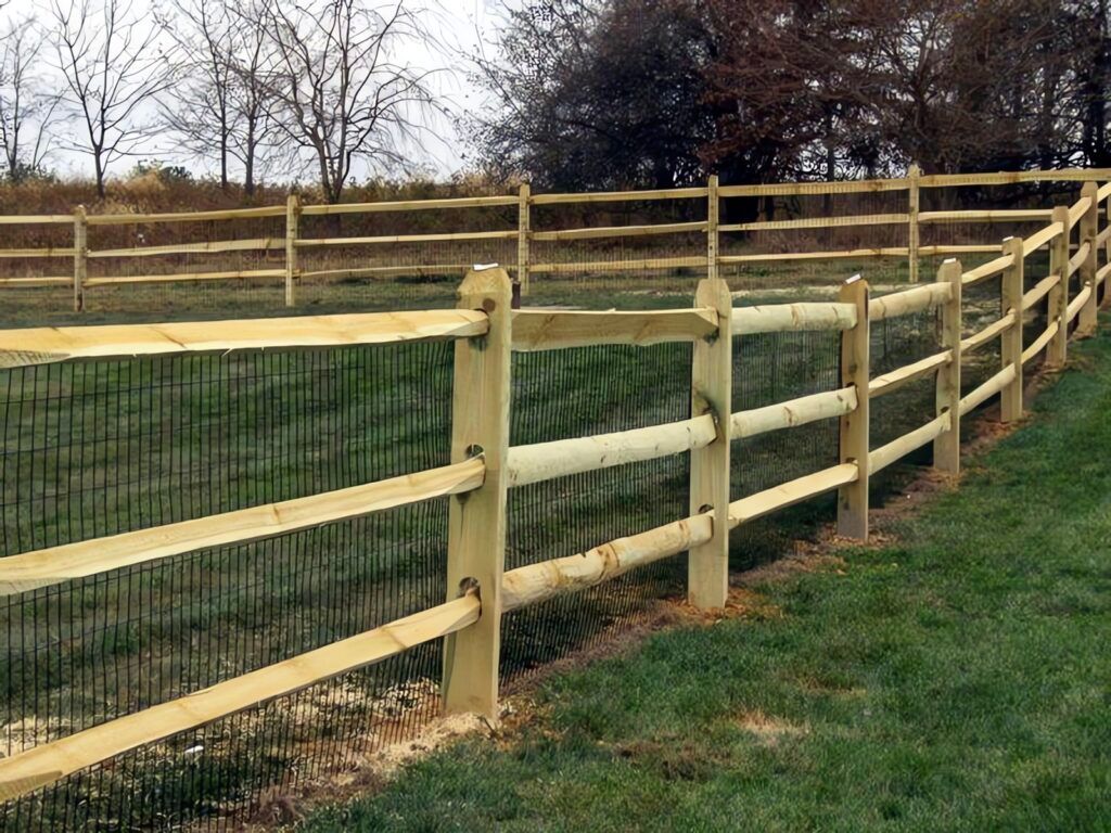 Wooden split-rail fence with black mesh backing surrounds a grassy area, in front of trees under a cloudy sky.