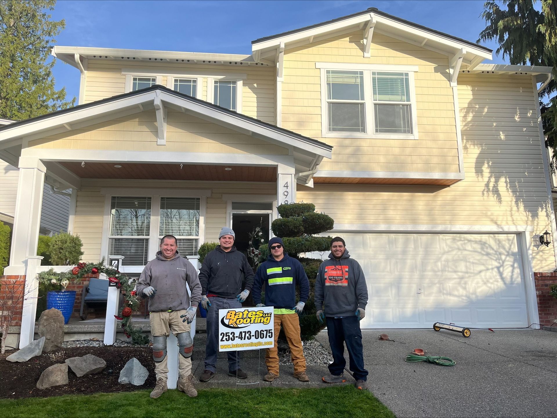 Four people stand in front of a yellow house, holding a company sign. They are on a gravel driveway.