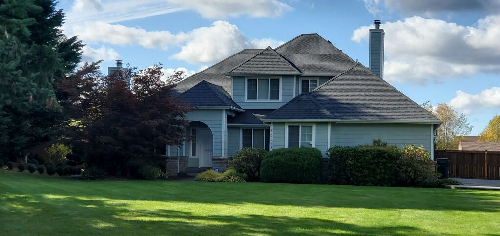 A two-story house with a green lawn, trees, and a cloudy blue sky.