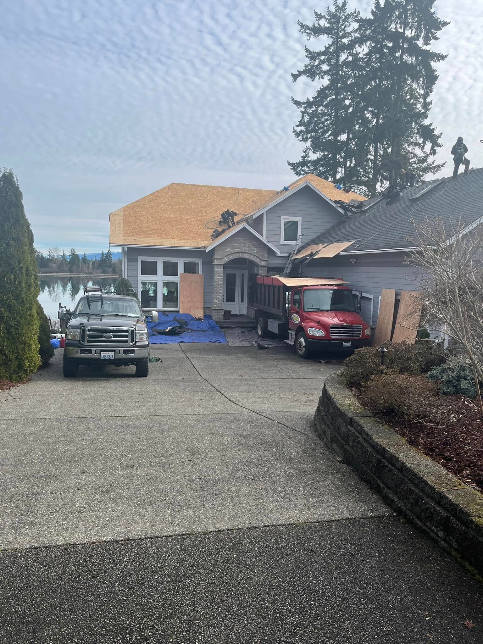 House under construction, driveway, trucks. Partial roof removed, debris on blue tarp. Cloudy day.