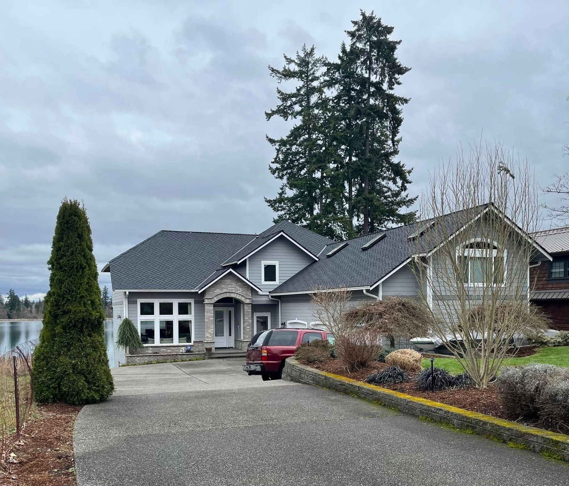 Gray house with stone accents, a red SUV in the driveway, and a lake view under a cloudy sky.