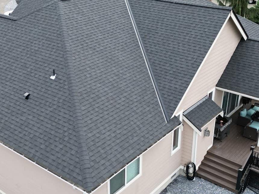 Gray shingled roof of a house with light tan siding and a brown deck.