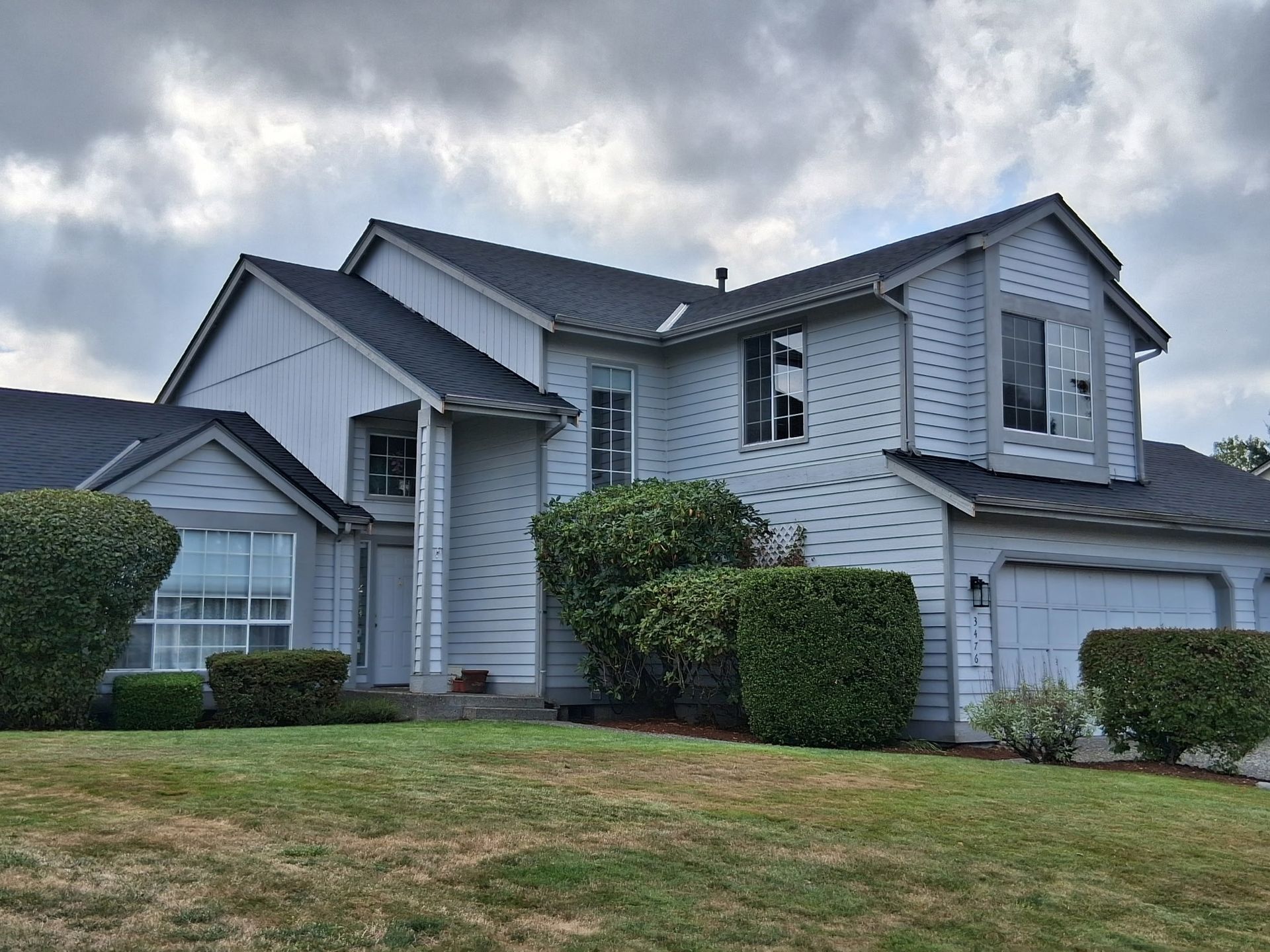Two-story light blue house with dark roof and green bushes in front on a grassy lawn under cloudy sky.