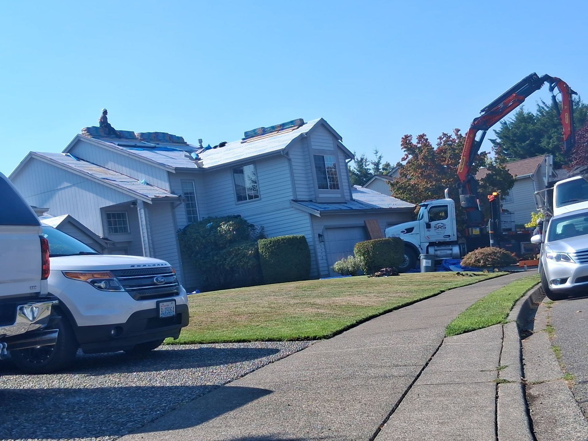 House with roof damage, crane removing debris; cars parked on street. Sunny day.