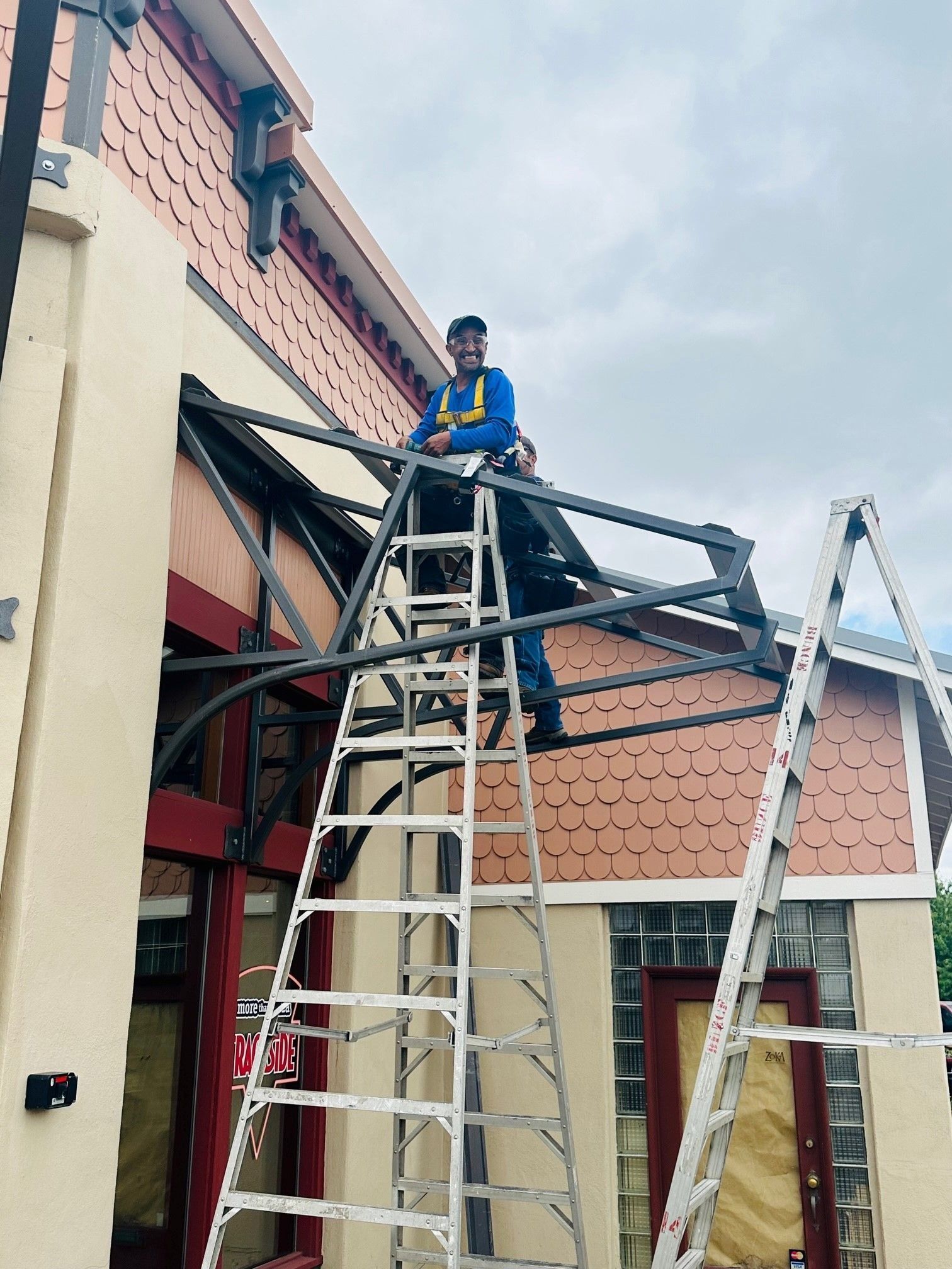 A worker on a ladder installs a metal awning frame on a building with red and beige facade.