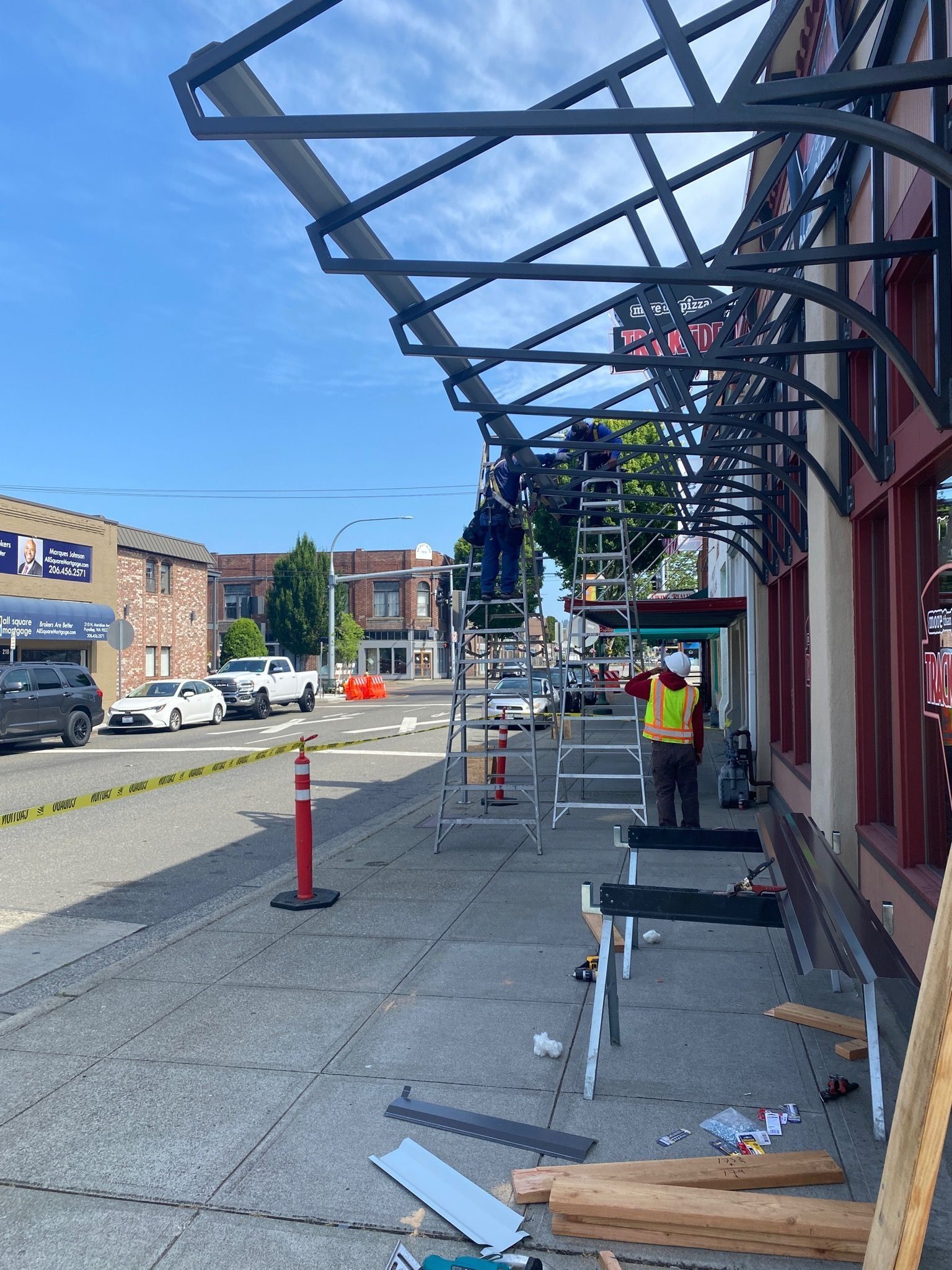 Workers installing a metal awning on a building facade next to a street with cars and other buildings.
