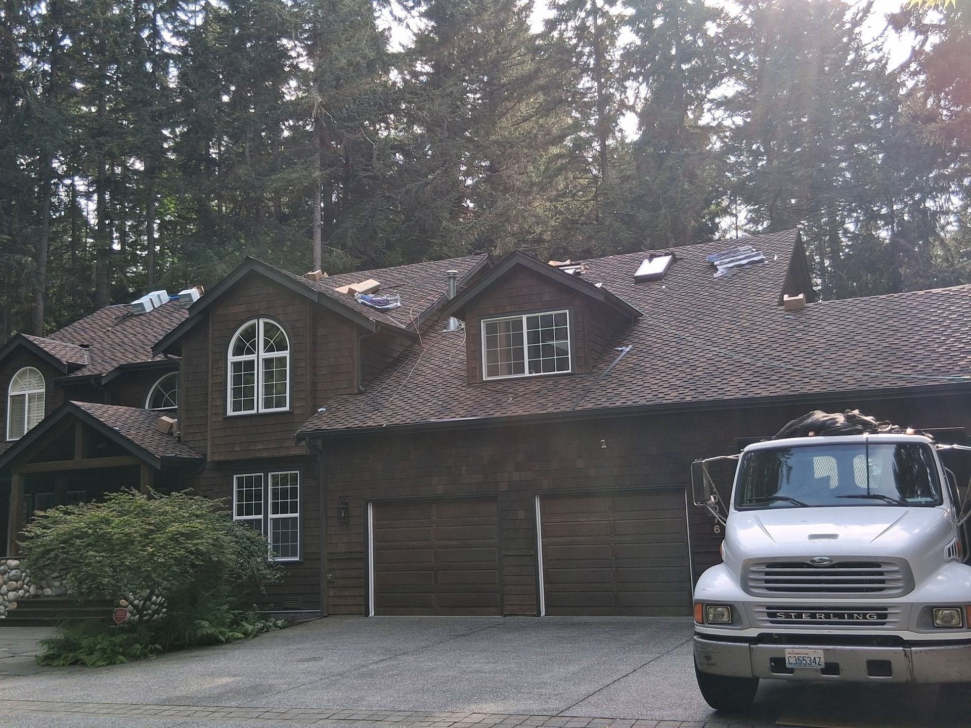 Brown house with a damaged roof, a truck parked in the driveway, and trees in the background.