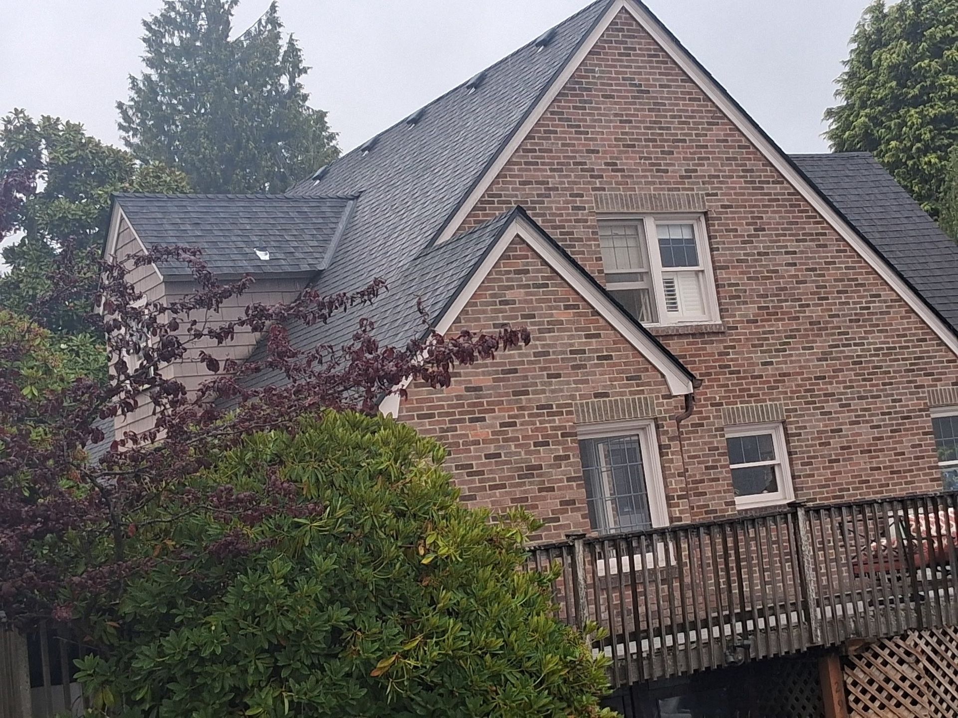 Brick house with dark roof and wooden deck, surrounded by trees. Cloudy sky.