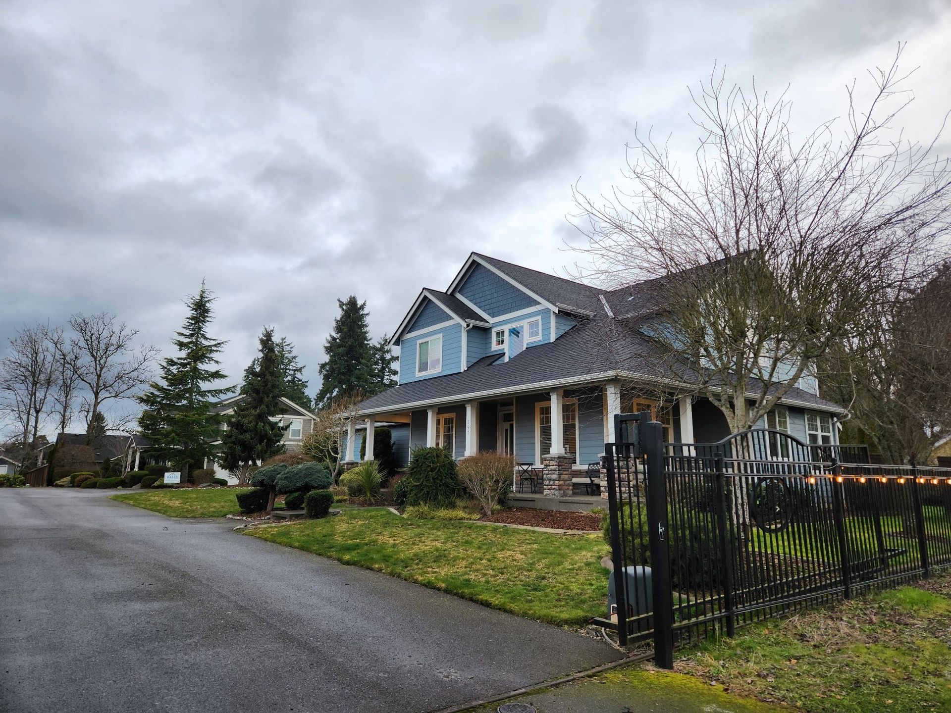 Blue house with a porch and dark gray roof on a cloudy day, with a wrought iron fence in the foreground.