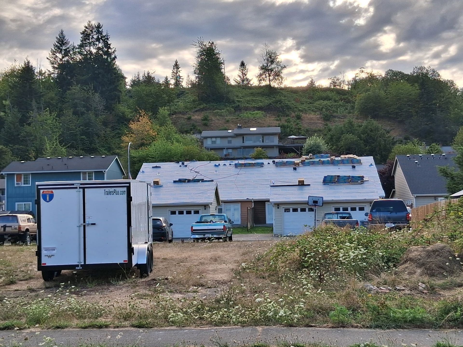House with roof in progress, surrounded by vehicles and vegetation, under a cloudy sky.