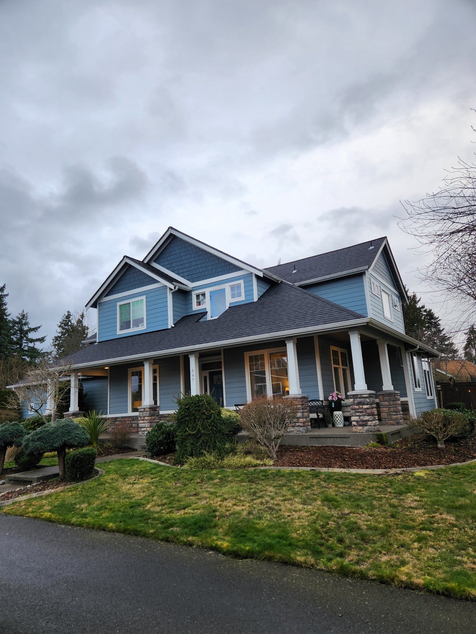 Two-story blue house with white trim, porch, and a dark roof under a cloudy sky.