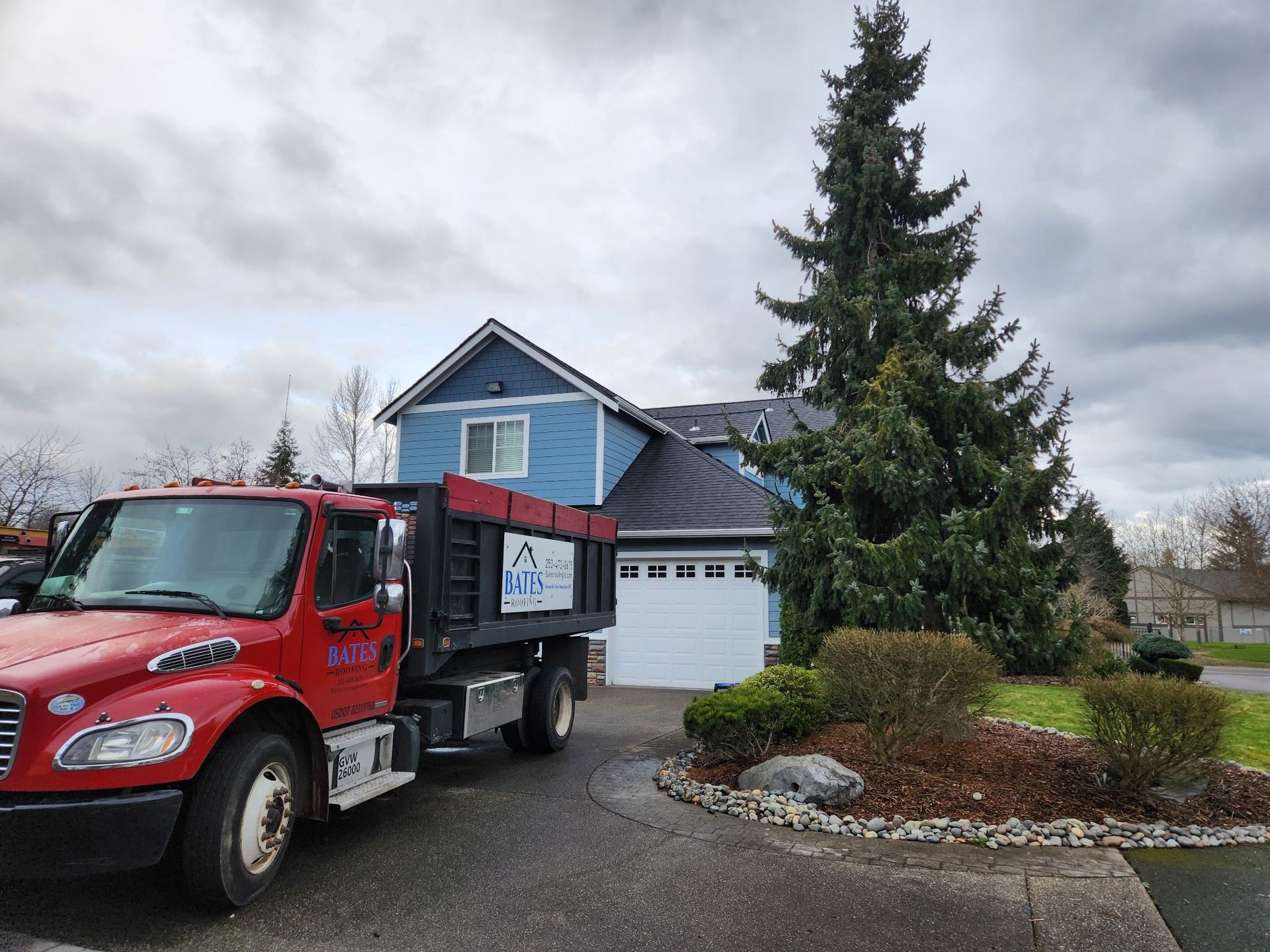 Red dump truck parked in front of a blue house with white garage doors, under cloudy skies.