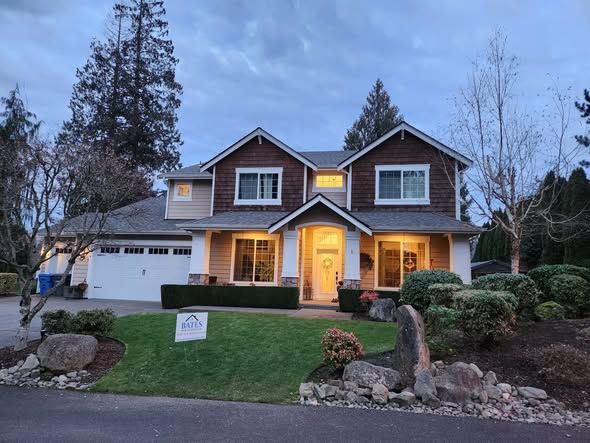 Two-story house with lit windows at dusk, front lawn, and landscaped yard with rocks and shrubs