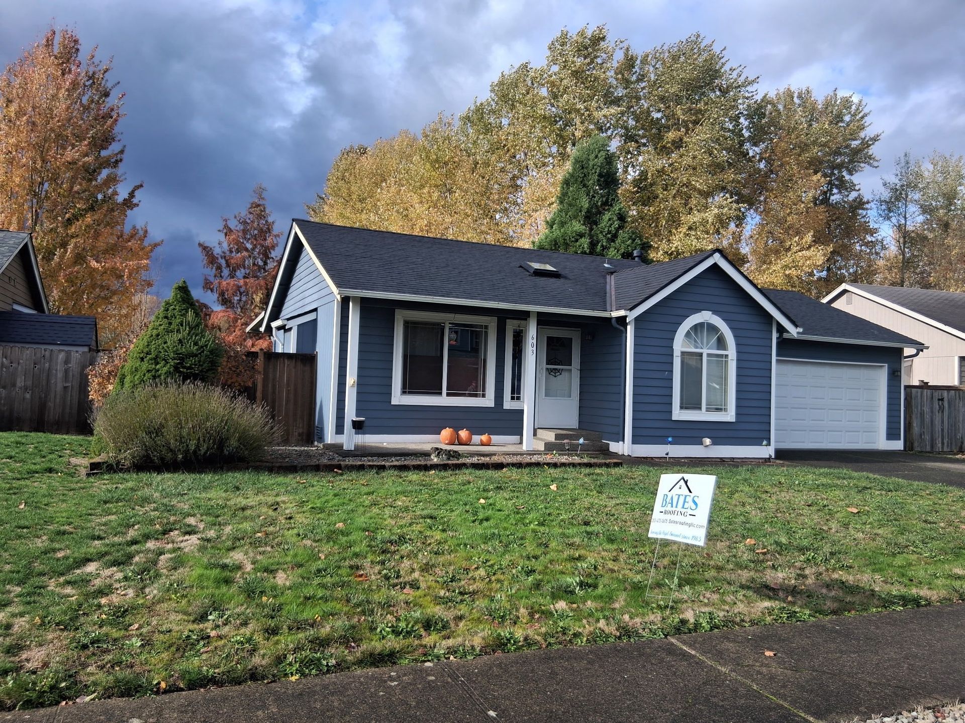 Blue house with a dark roof and white trim in front of autumn trees, overcast sky.