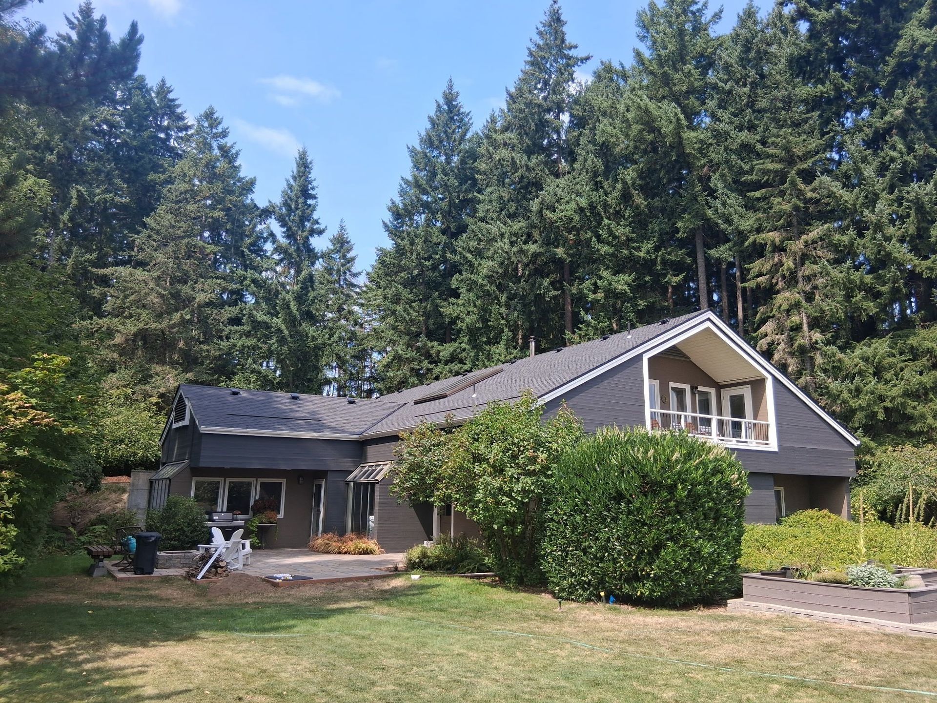 House with dark siding and a damaged roof, set against tall evergreen trees and a blue sky.