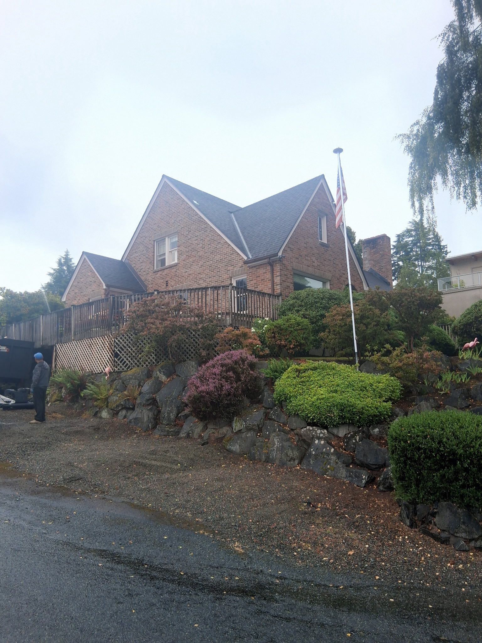 Brick house with sloped roof, surrounded by landscaping and a stone retaining wall. A person stands near a vehicle.