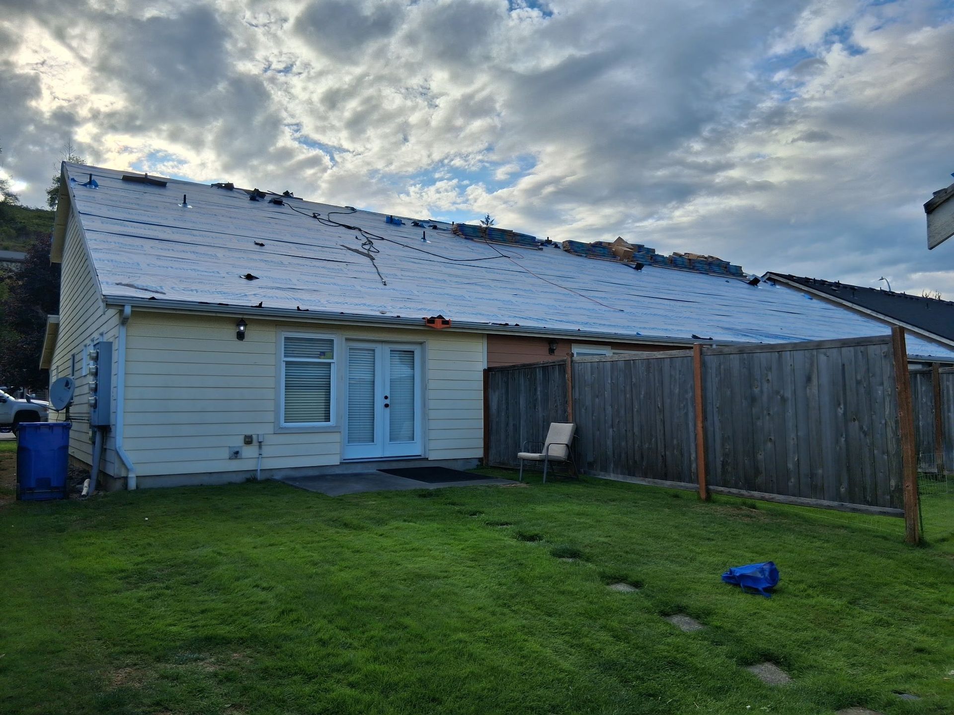 House with a partially replaced roof. White siding, wooden fence, cloudy sky, green grass.