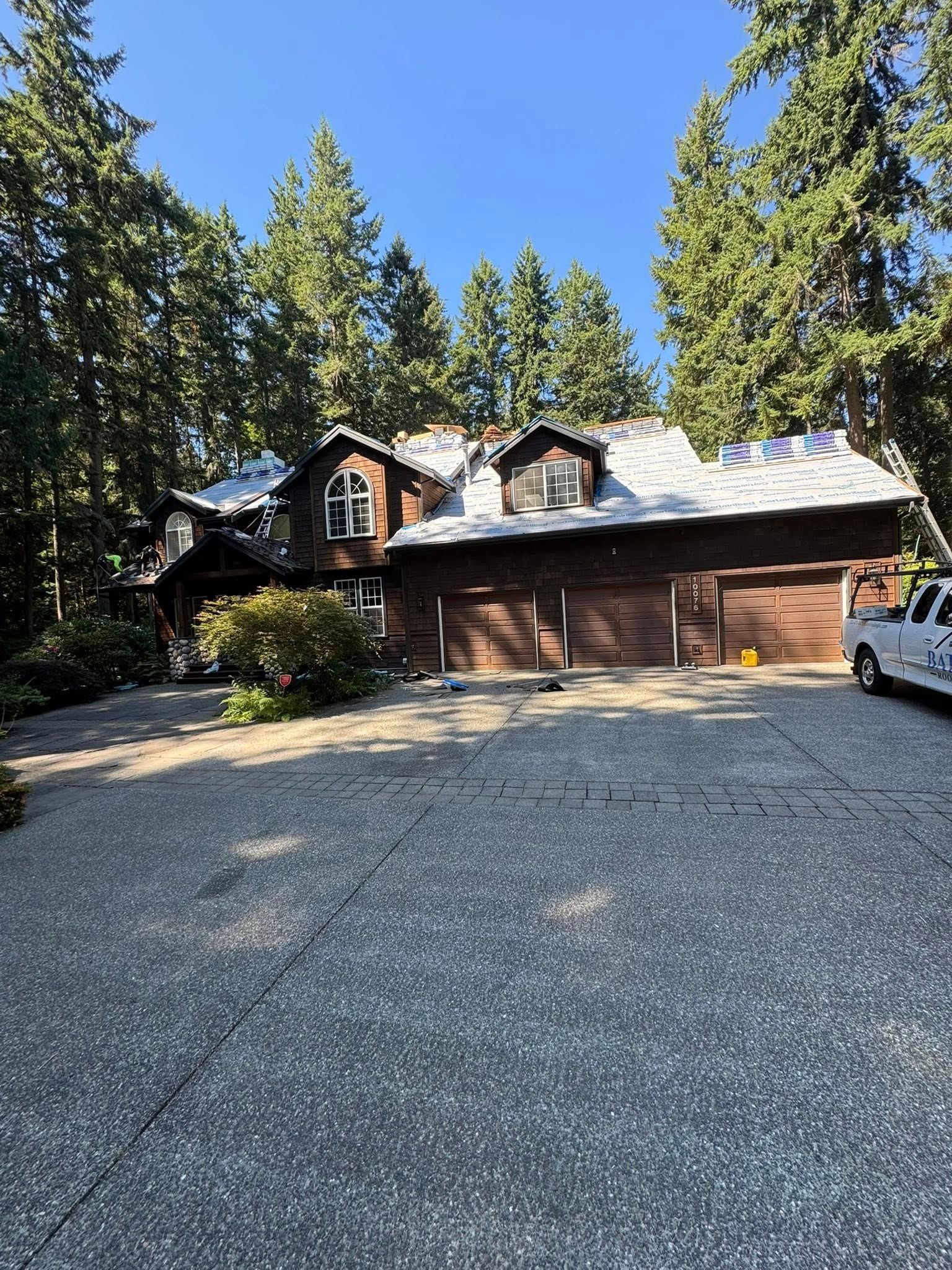 Brown wooden house with three garage doors and a gravel driveway, surrounded by trees.