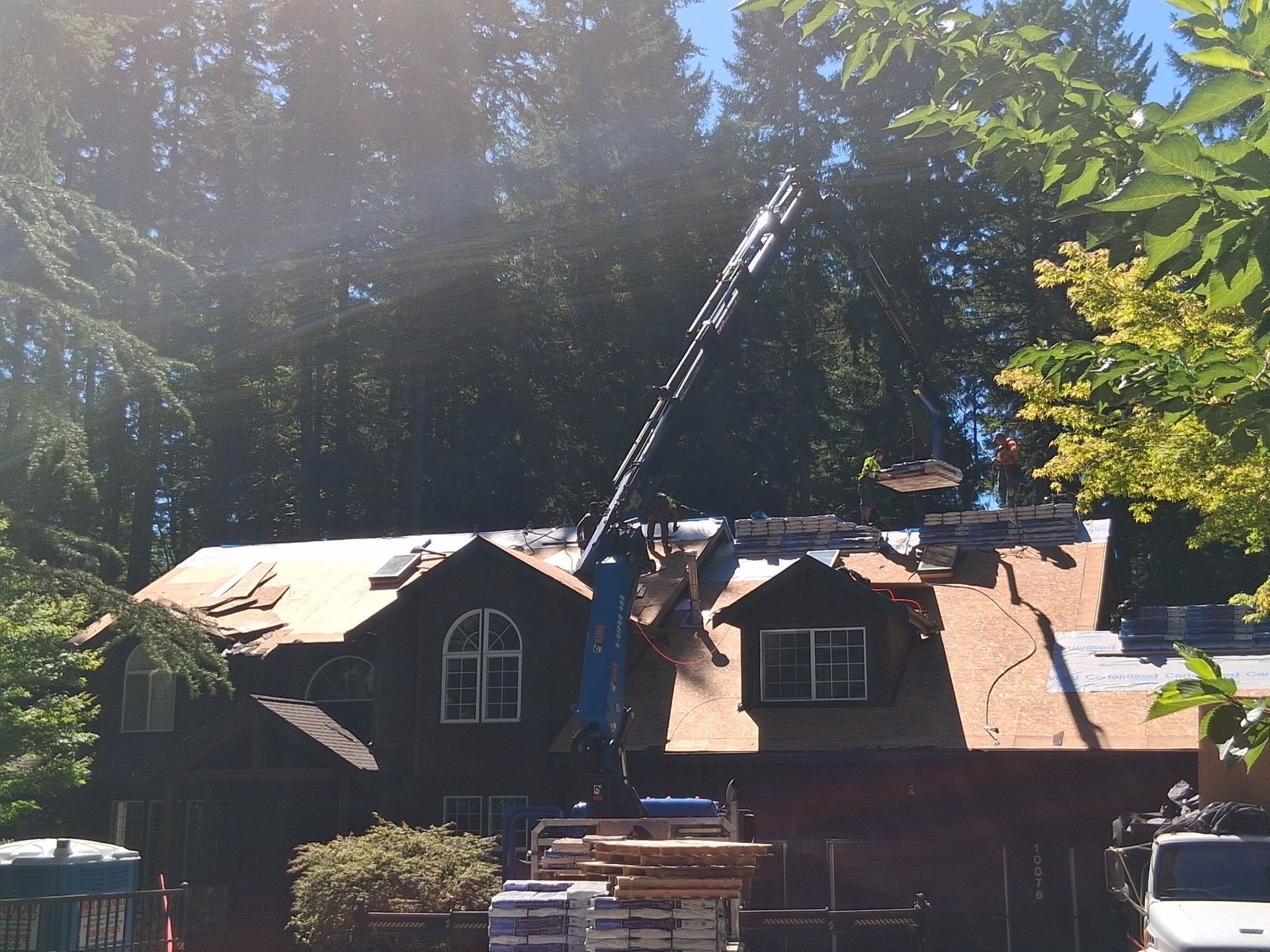 House with roof being repaired. Crane lifting materials; green trees in the background, sunny day.