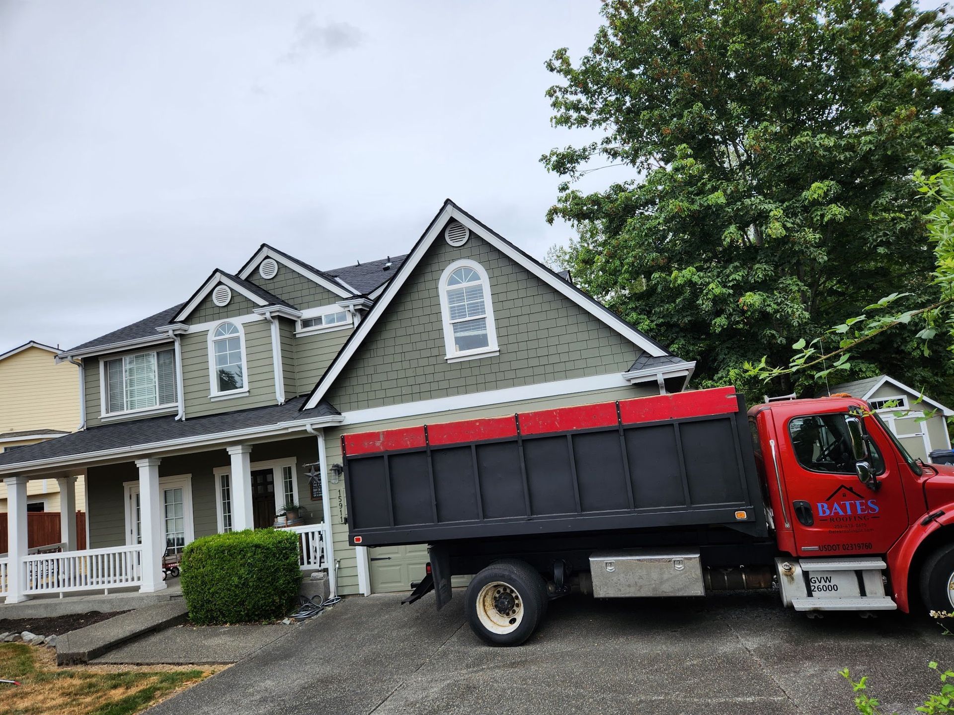 Red dump truck parked in front of a green two-story house, ready to load debris. Overcast sky.