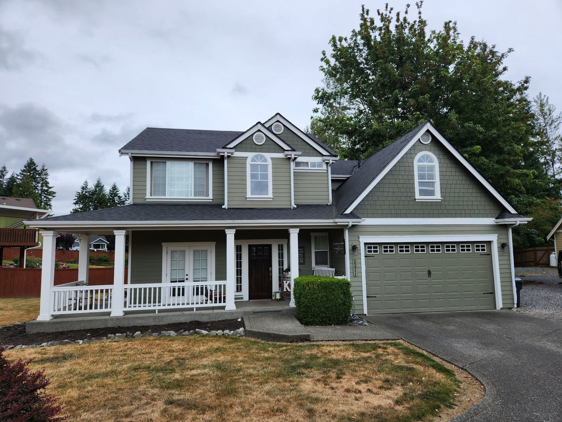 Green two-story house with porch, white trim, and attached garage on a cloudy day.