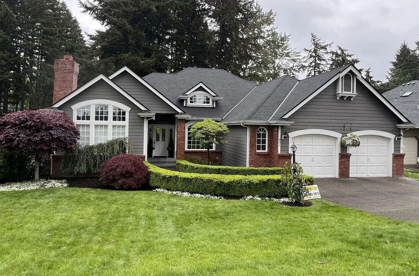 Gray house with brick accents, dormers, and a two-car garage. Landscaping includes green lawn and hedges.