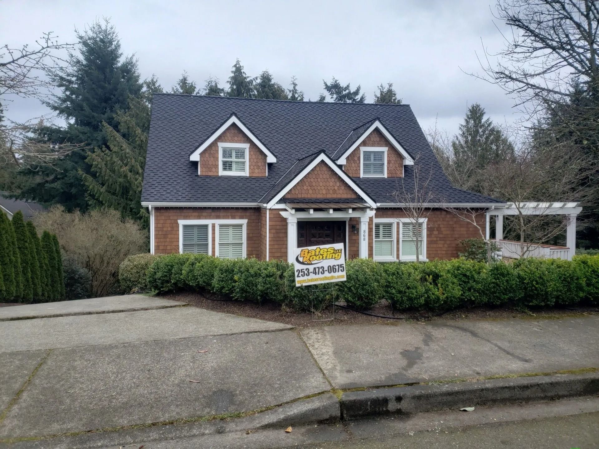 Brown house with dark roof and dormers, surrounded by green shrubs, on a gray day.