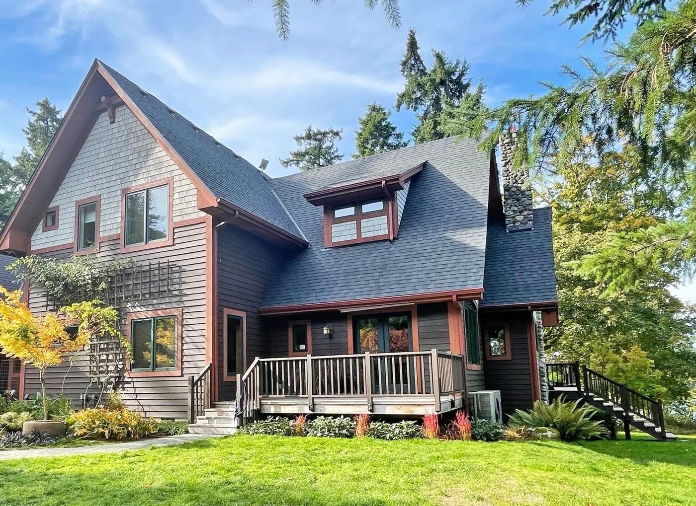 A wood-sided house with a dark roof and a porch, surrounded by trees and green grass.