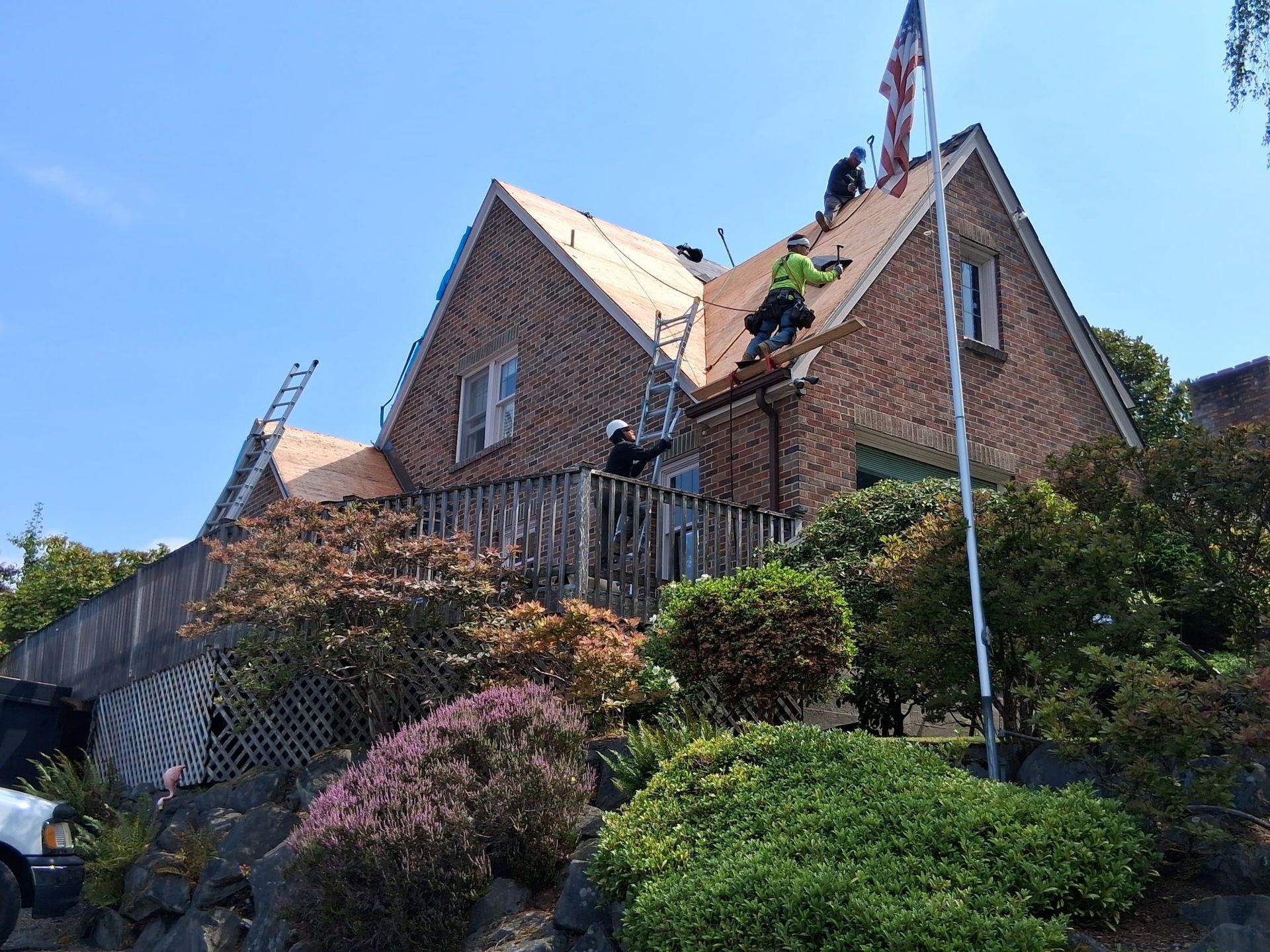 Roofers working on a brick house roof with scaffolding; American flag flies.