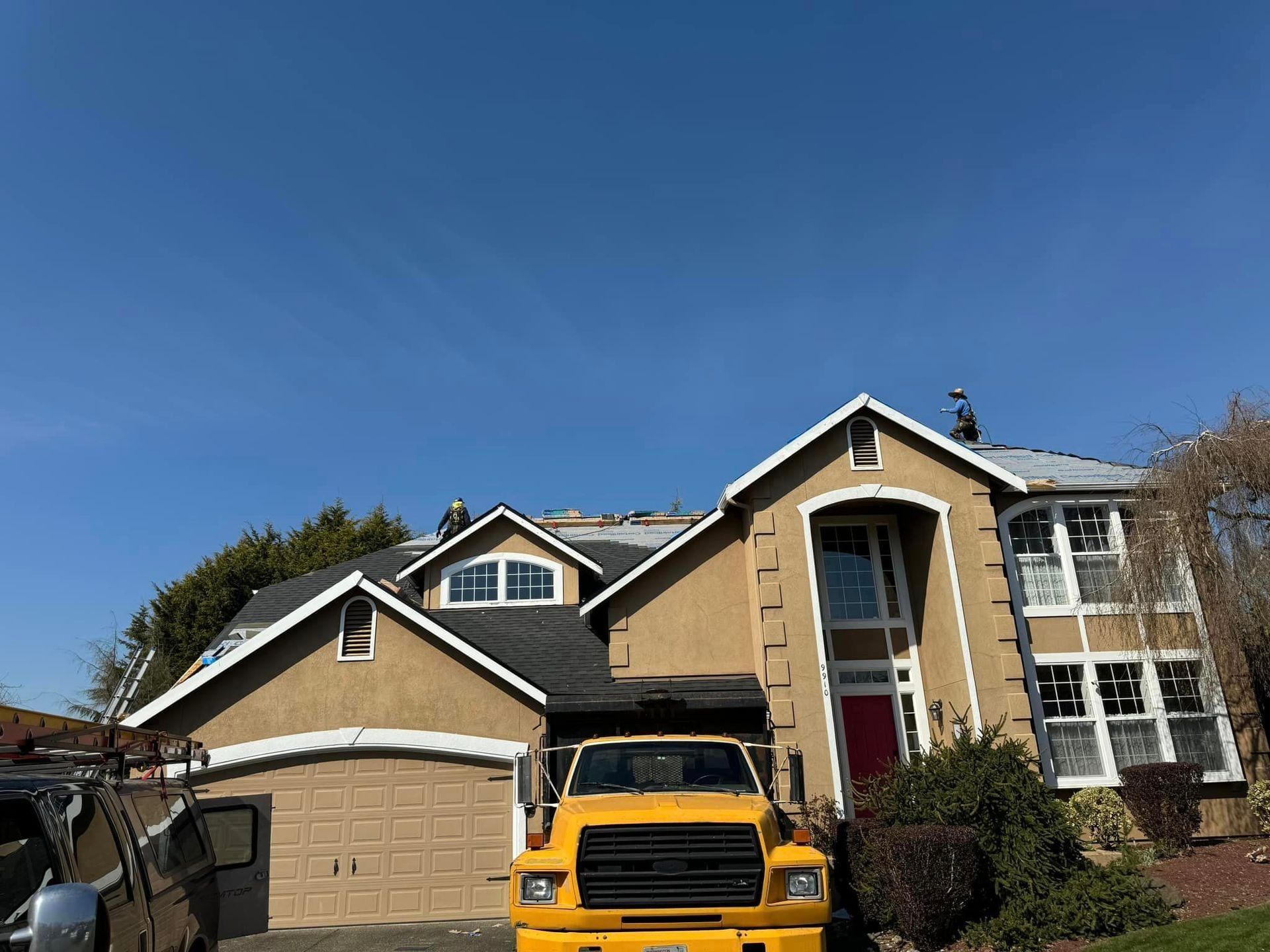 Yellow dump truck in front of a tan two-story house with a partially removed roof. Blue sky.