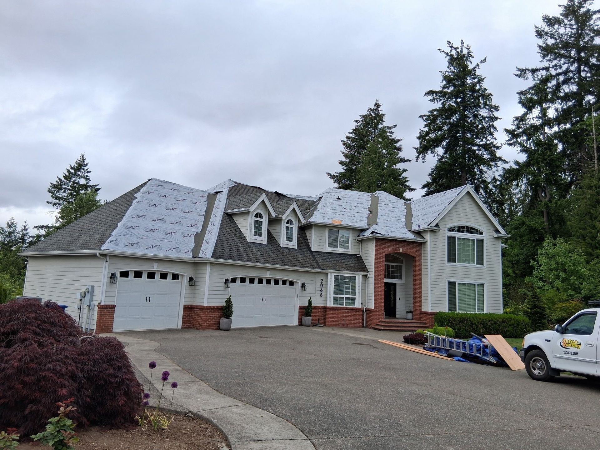 House with a partially replaced roof; white exterior, three-car garage, gray driveway, truck parked on right.