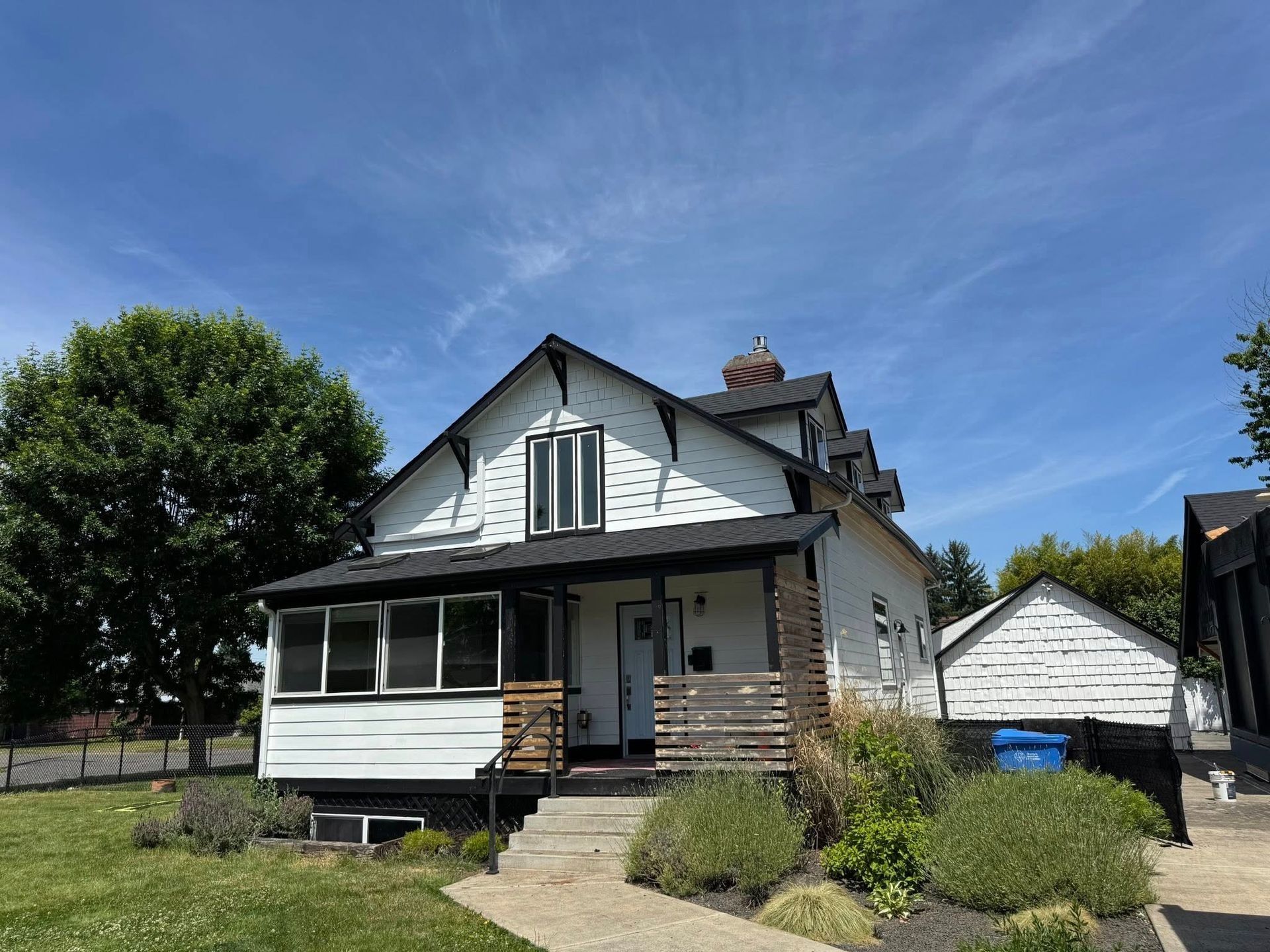 White two-story house with black roof and trim, porch with wooden slats, bushes, and a blue sky.