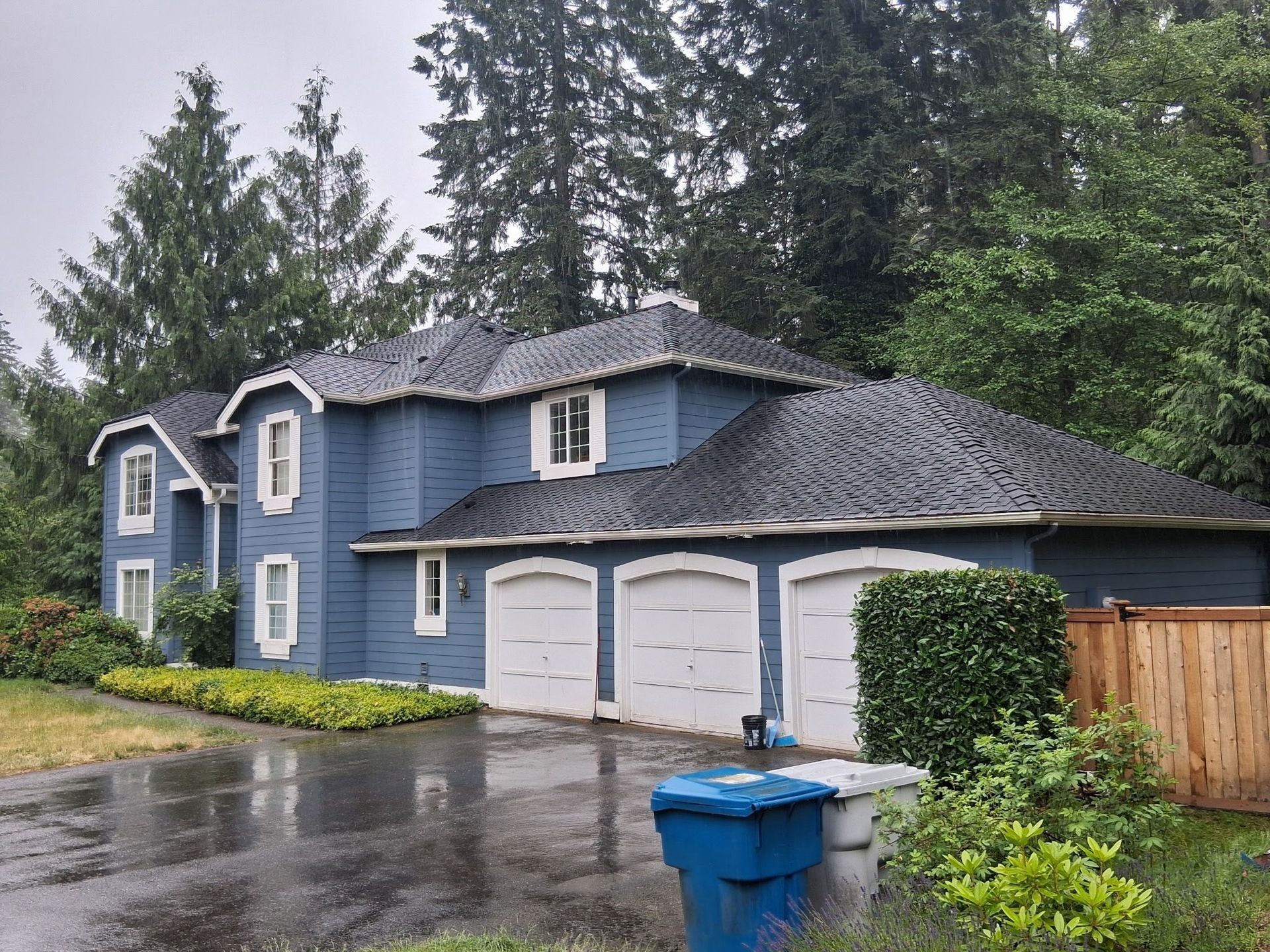 Blue house with three garage doors; wet driveway, overcast sky, trees.