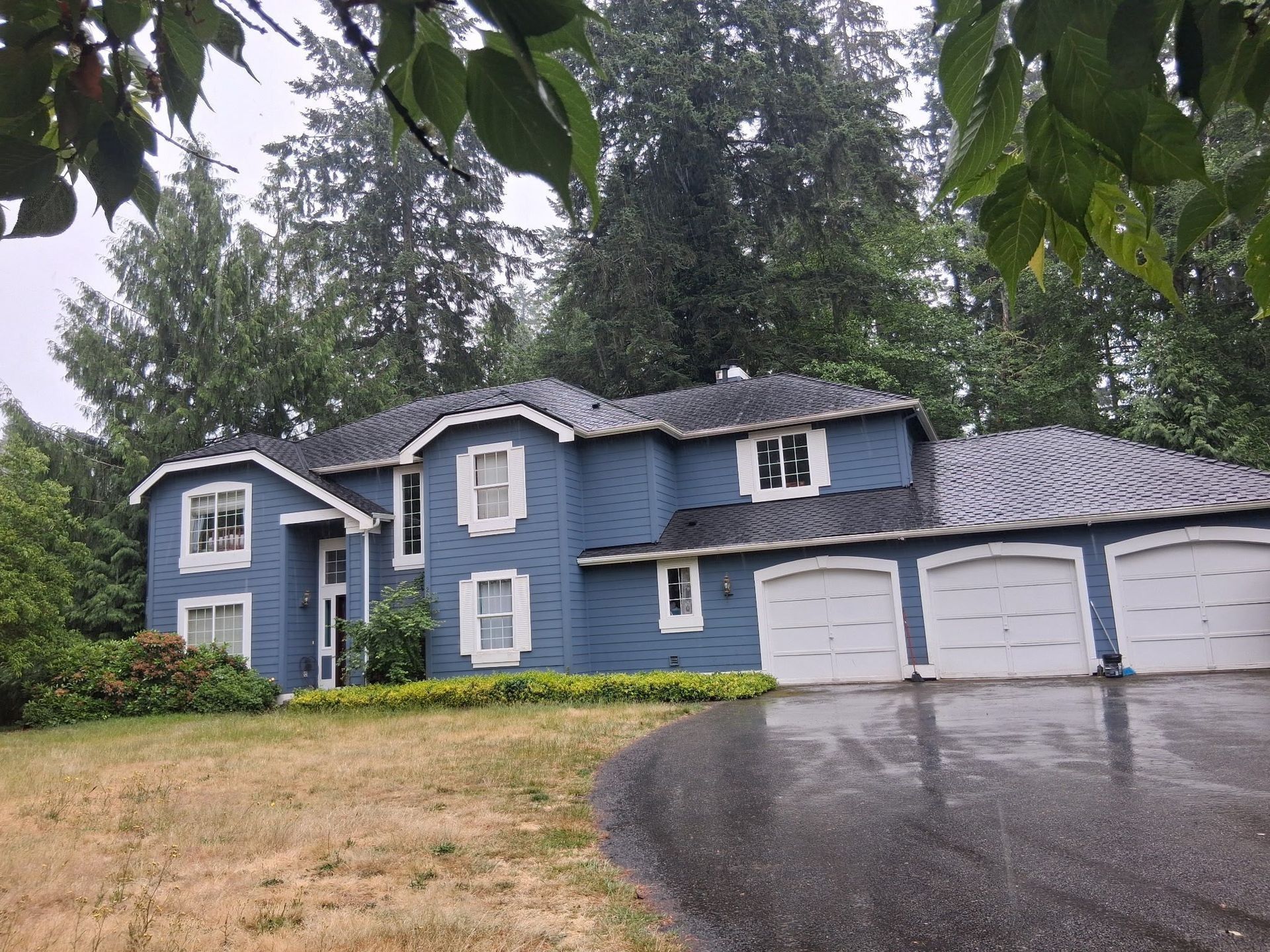Blue two-story house with white trim, three-car garage, and a wet driveway surrounded by trees.