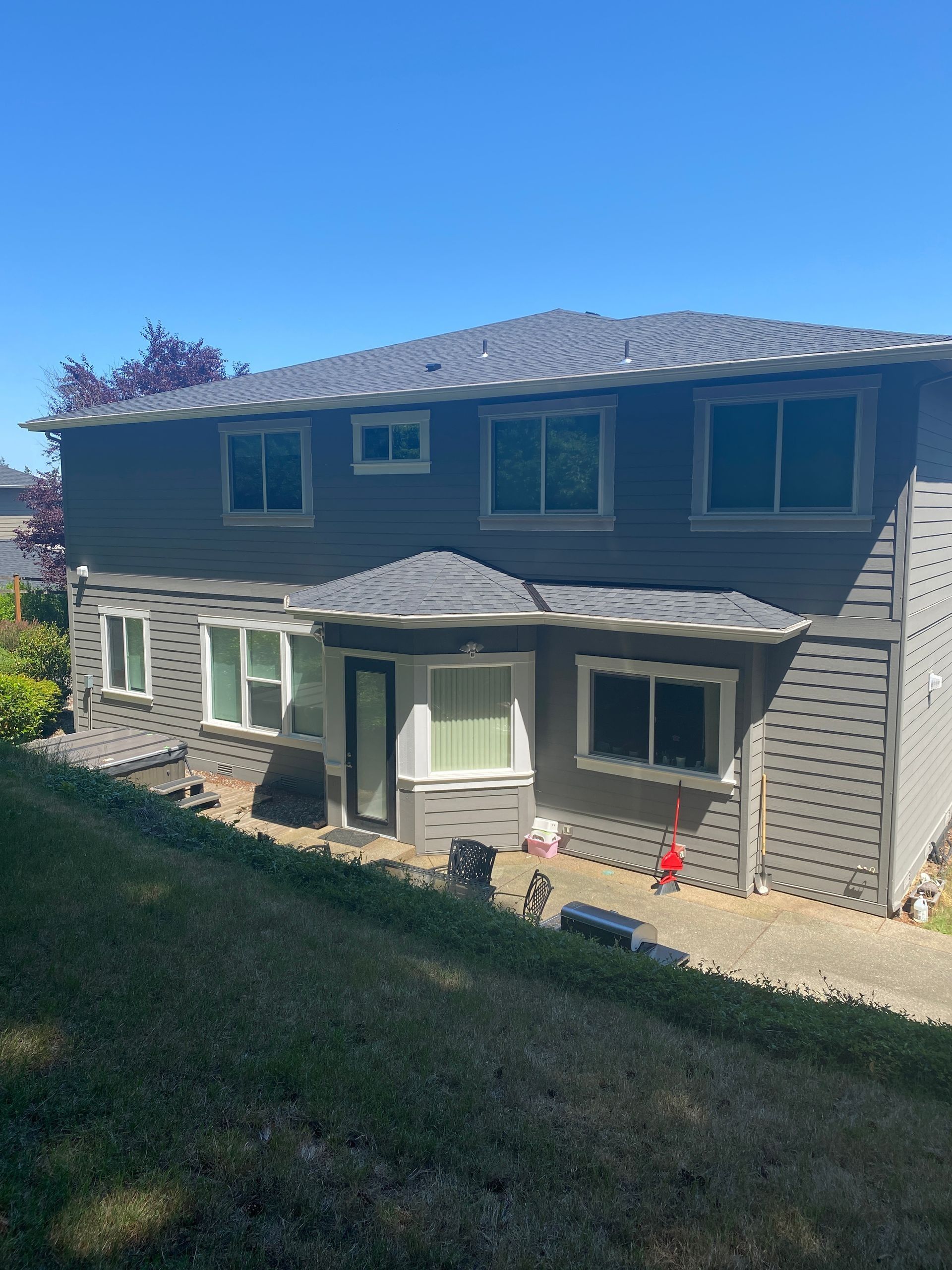 Gray two-story house with dark roof and windows, set on a grassy hill under a blue sky.