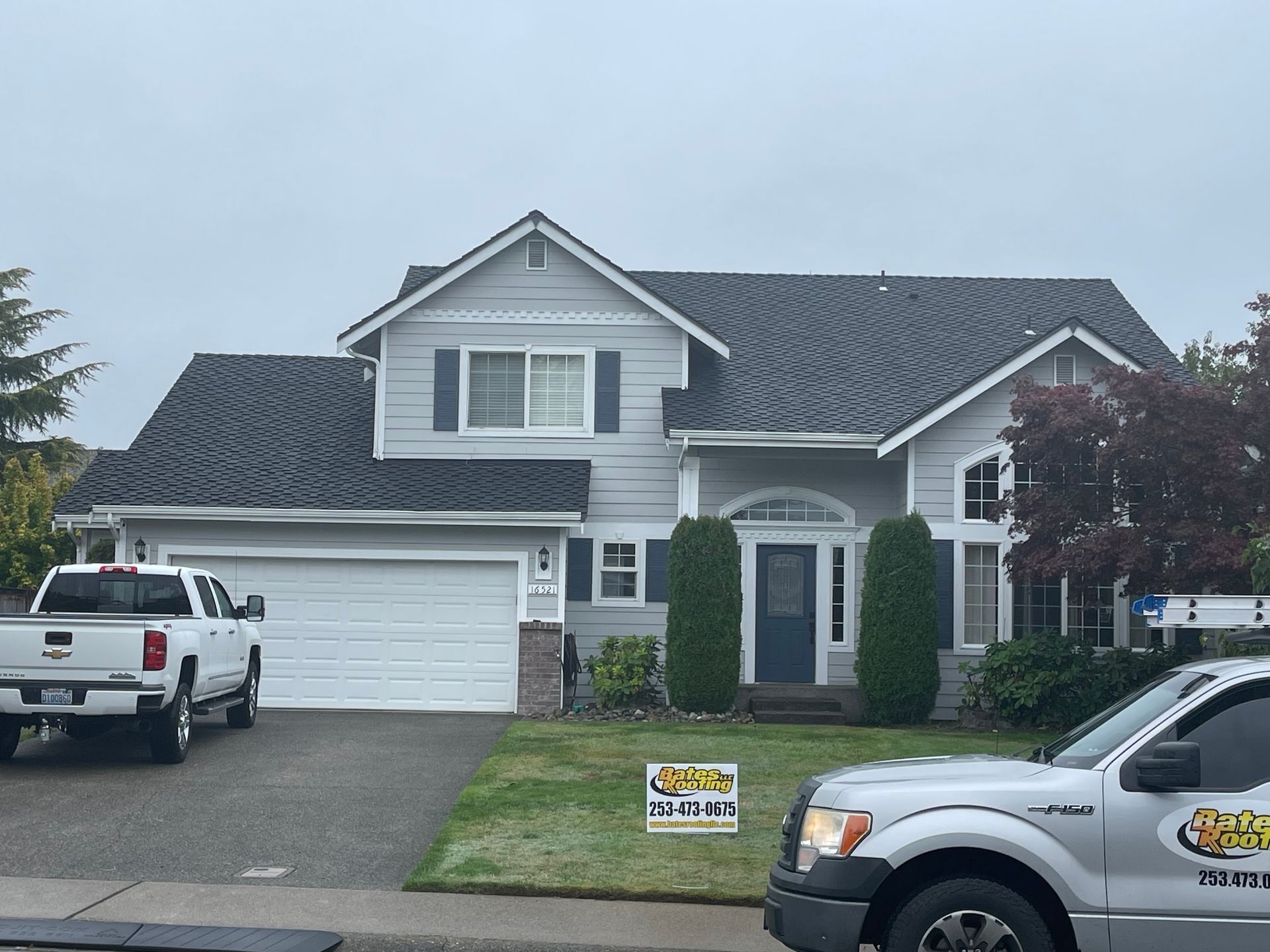 Two-story house with gray siding, dark roof. White garage door, truck parked in driveway. Gray sky.