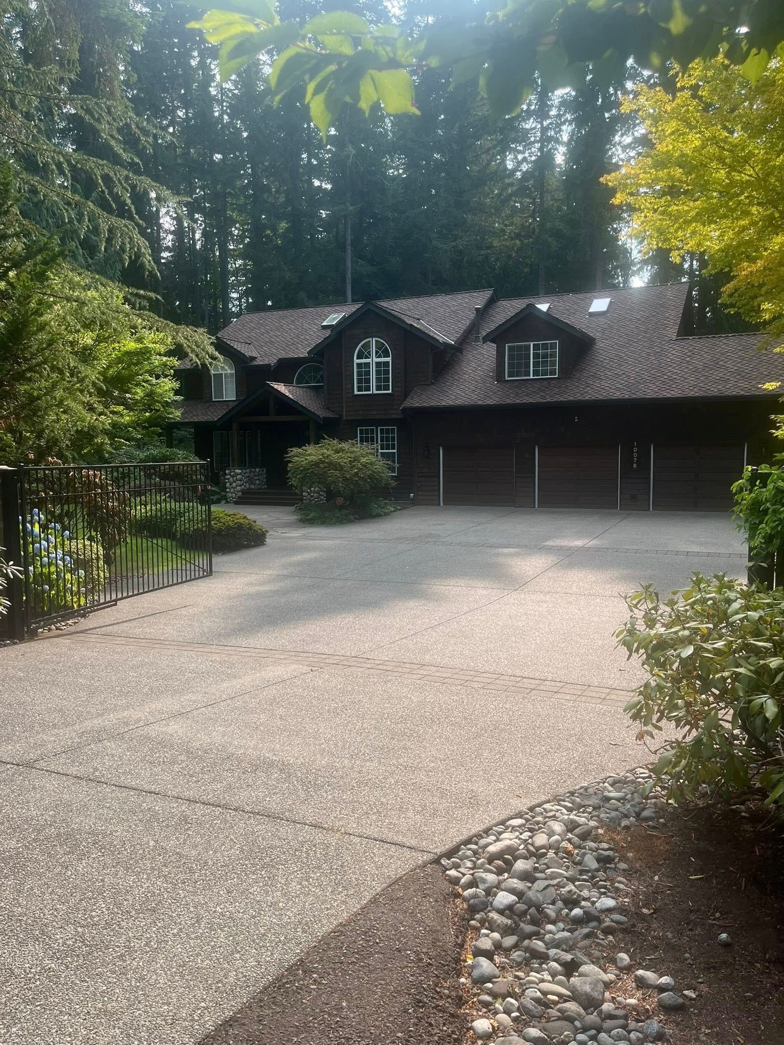Brown wooden house with a gravel driveway, surrounded by trees and bushes under a bright sky.