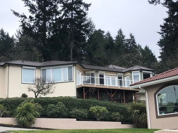 A light-colored house with a deck sits atop a retaining wall, overlooking a road and trees.