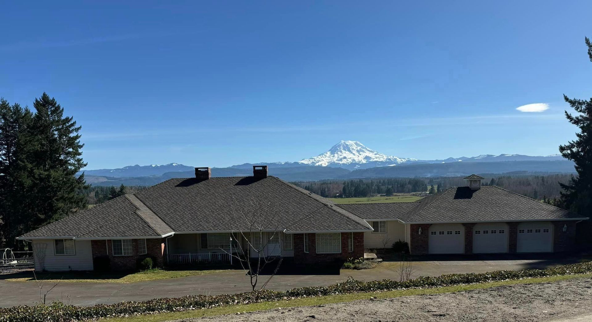 House with a gray shingled roof, trees, and snow-capped mountain in the background under a blue sky.