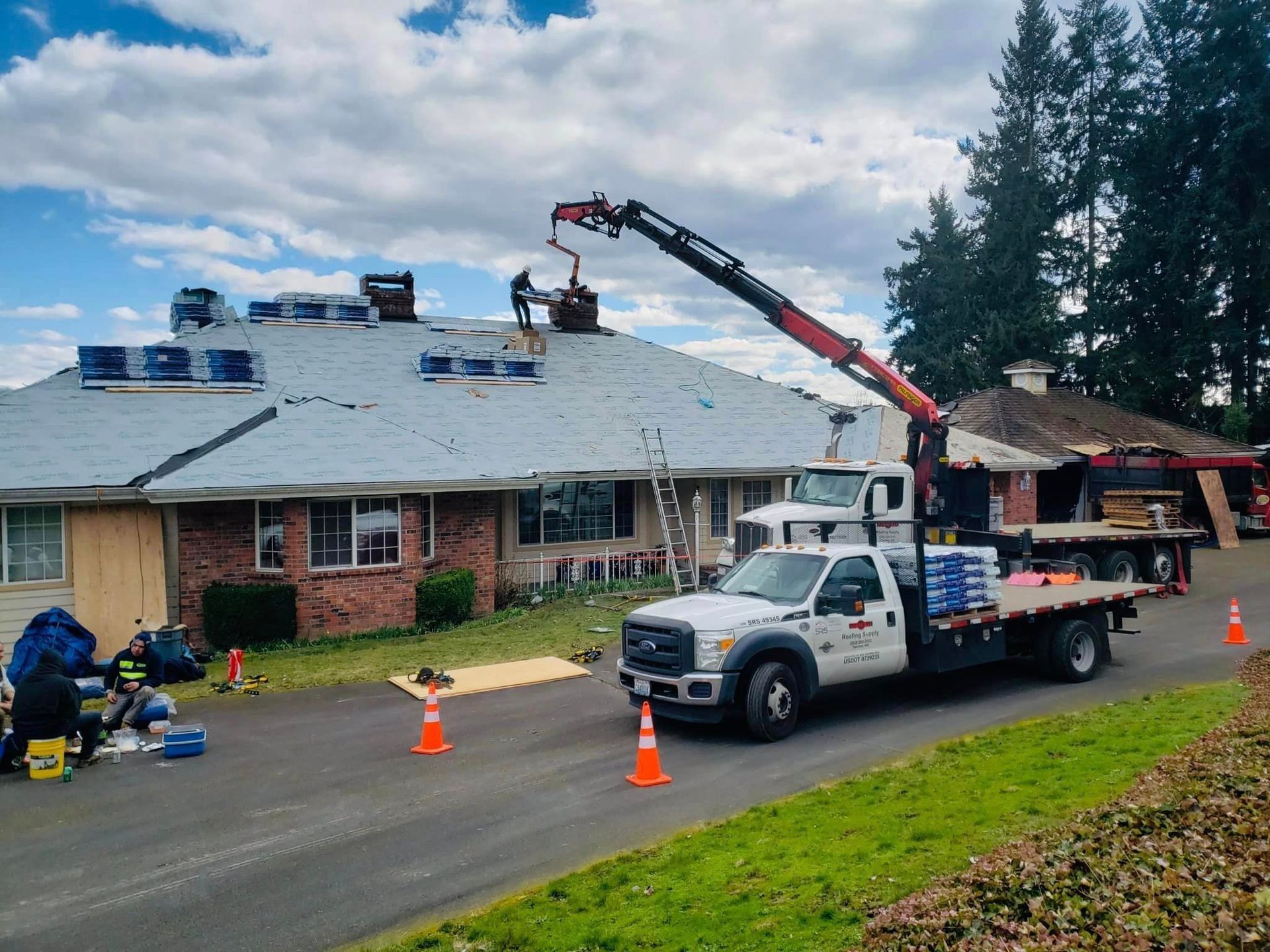Truck with crane on residential roof, workers repairing chimneys, materials on roof and ground.