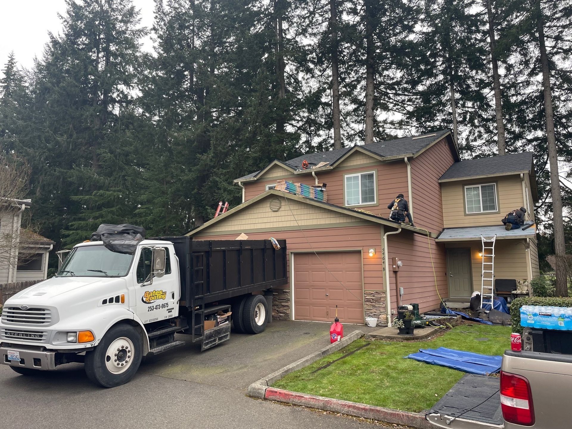 Truck and workers on a roof, preparing to work on a house's roof in front of trees and a green lawn.
