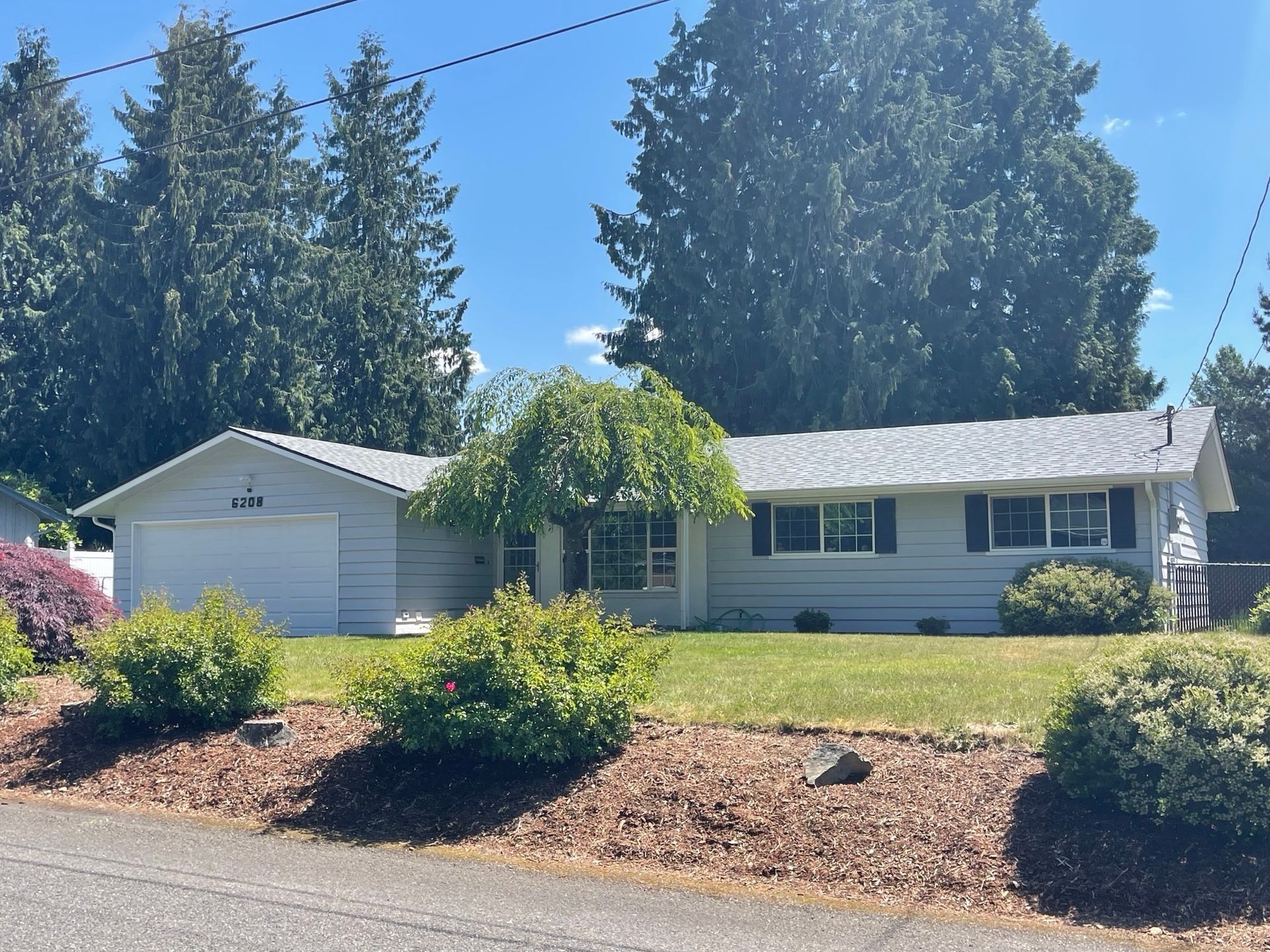 Light blue single-story house with a white garage. Green lawn, trees in the background, blue sky.