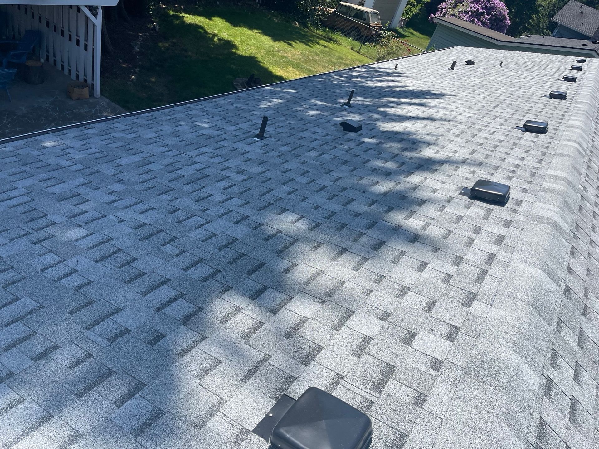 Gray asphalt shingle roof with vents and pipes, angled view from above in daylight.